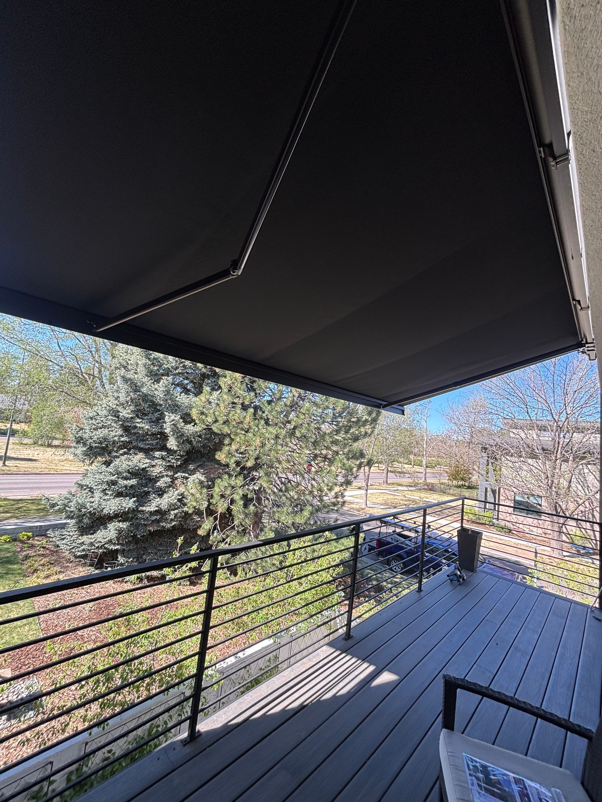 Covered balcony with black railing, wooden floorboards, and a view of trees and blue sky.