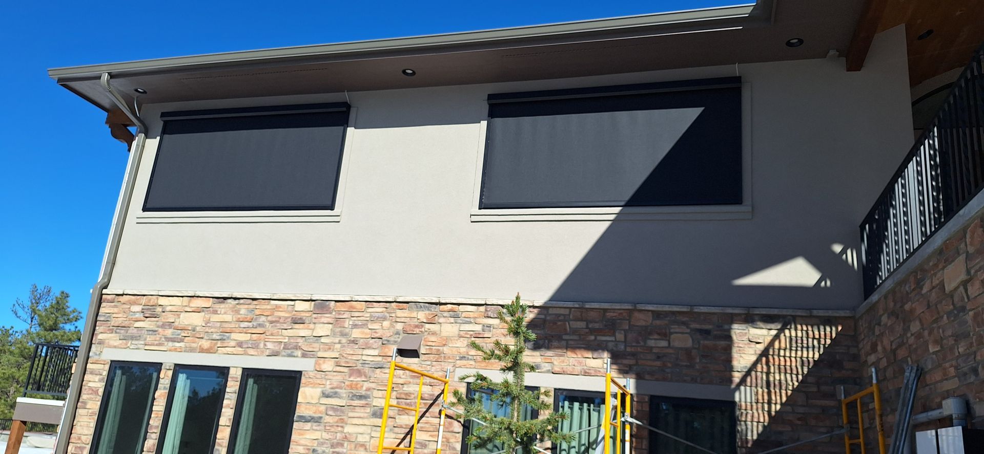 Modern two-story house exterior with beige walls, stone accents, and dark window shades in bright sunlight