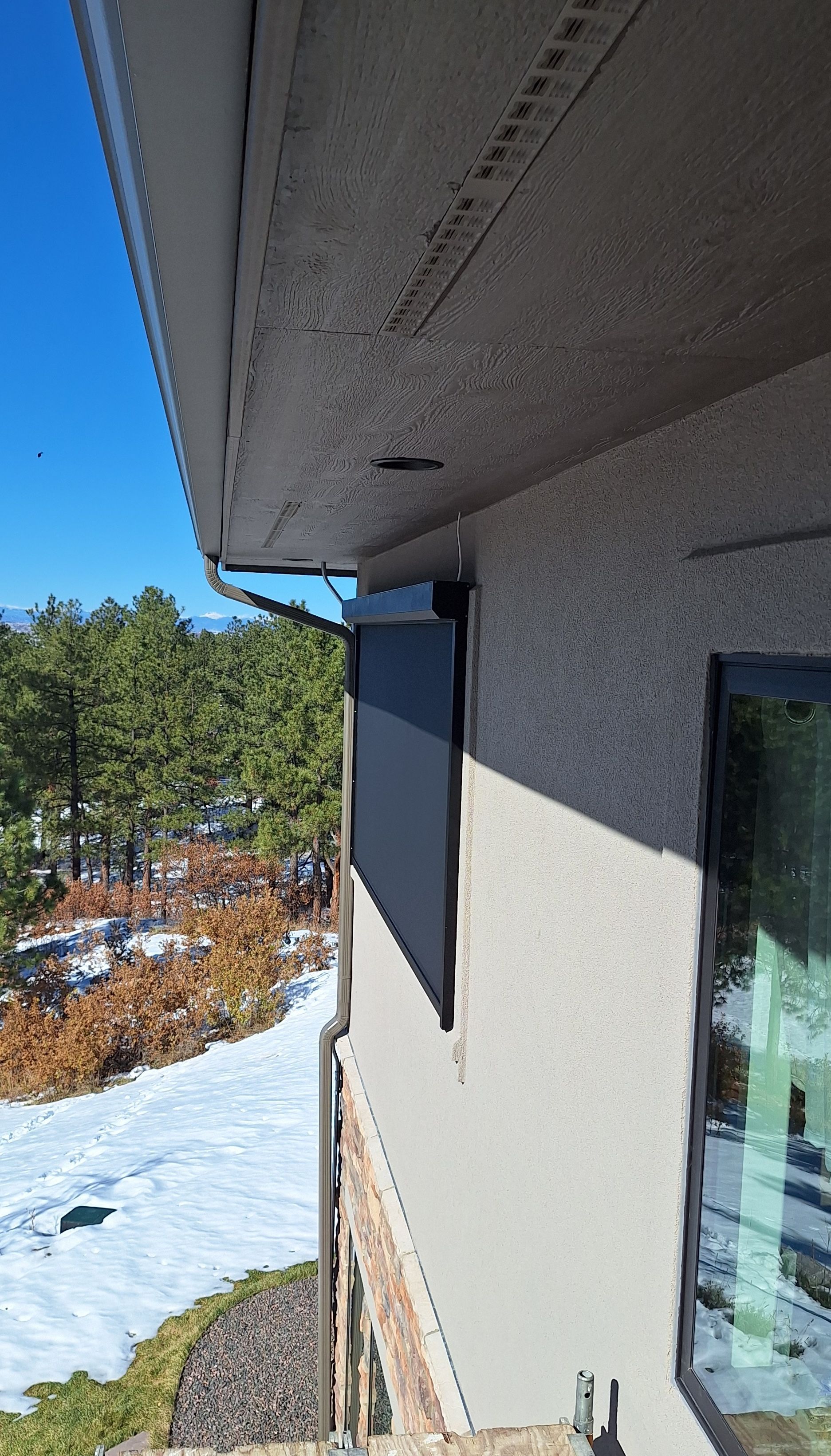 Snowy backyard beside house with white eaves, window, and trees under a clear blue sky