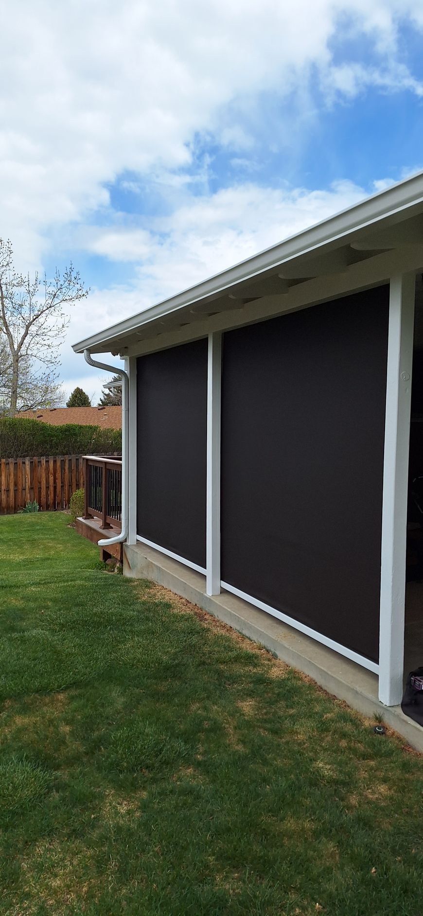Covered patio with dark roll-down shades beside a grassy yard and wooden fence