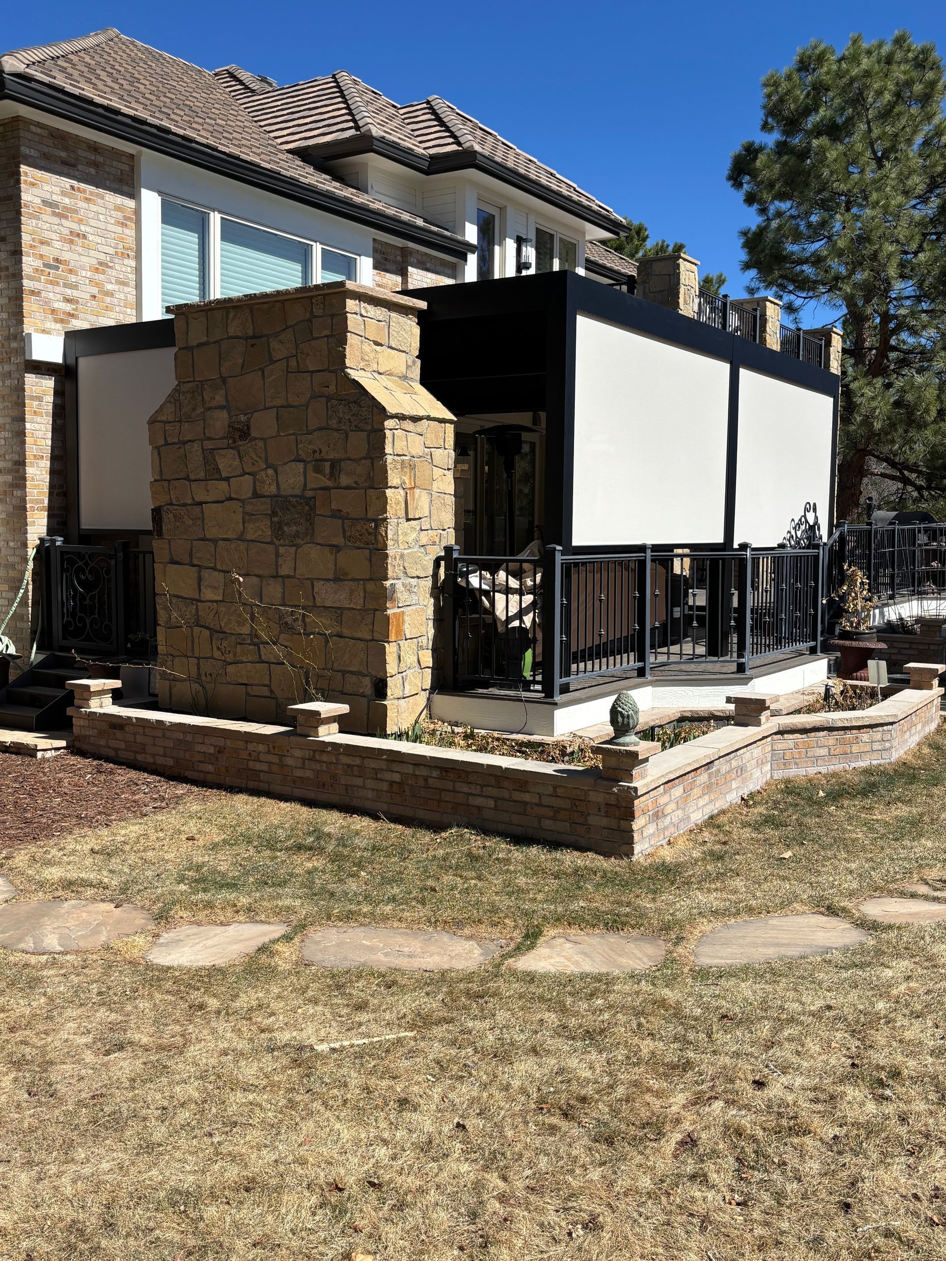 Patio extension with stone pillar, black railing, and white panels beside a house in a grassy backyard