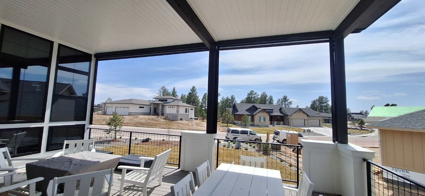 Covered patio with white tables and chairs, overlooking a neighborhood and blue sky beyond black support beams.