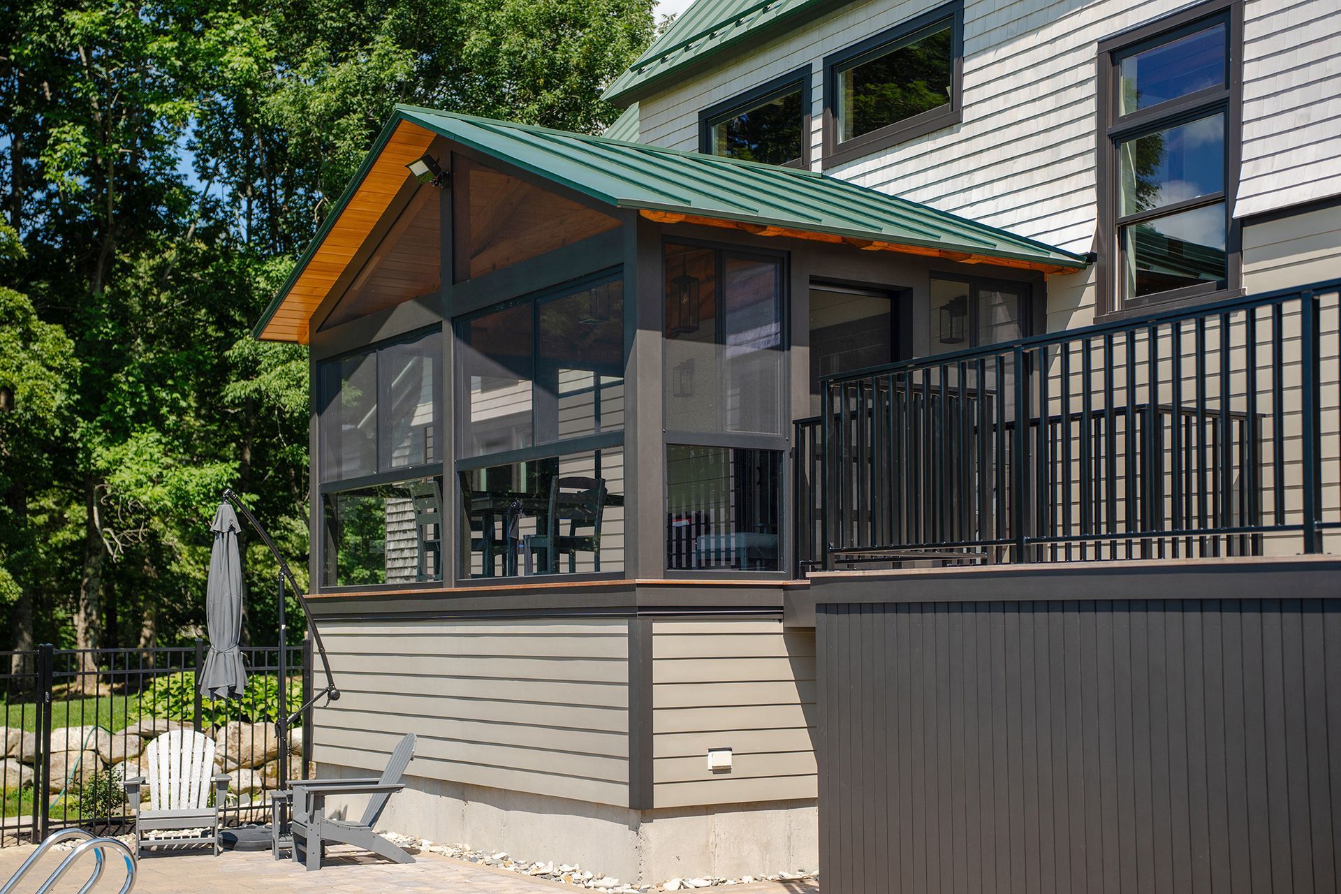 Screened porch and deck on a house with gray siding, black railings, and a green roof in a wooded yard