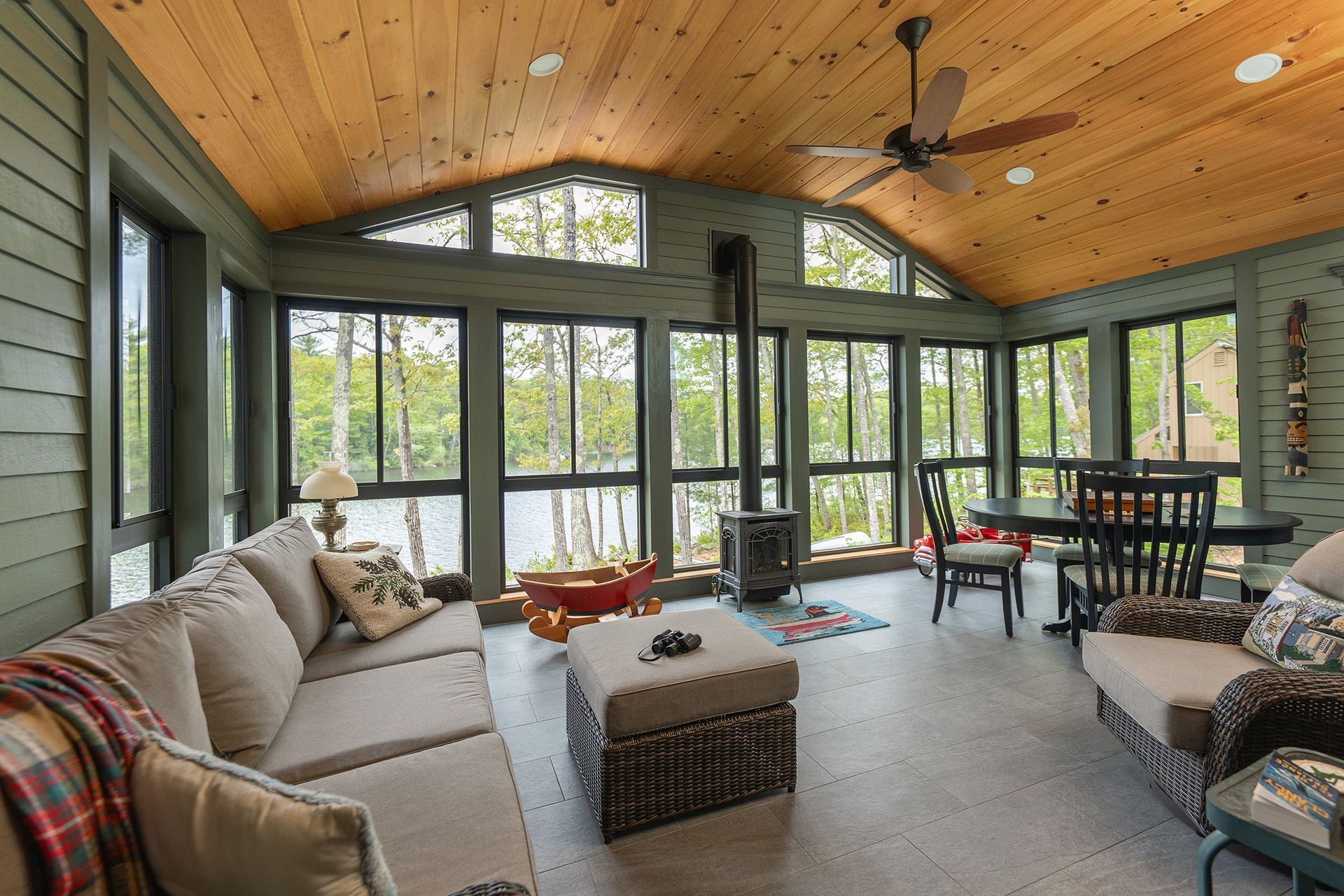 Bright sunroom with vaulted wood ceiling, large windows, sectional sofa, and dining table overlooking greenery