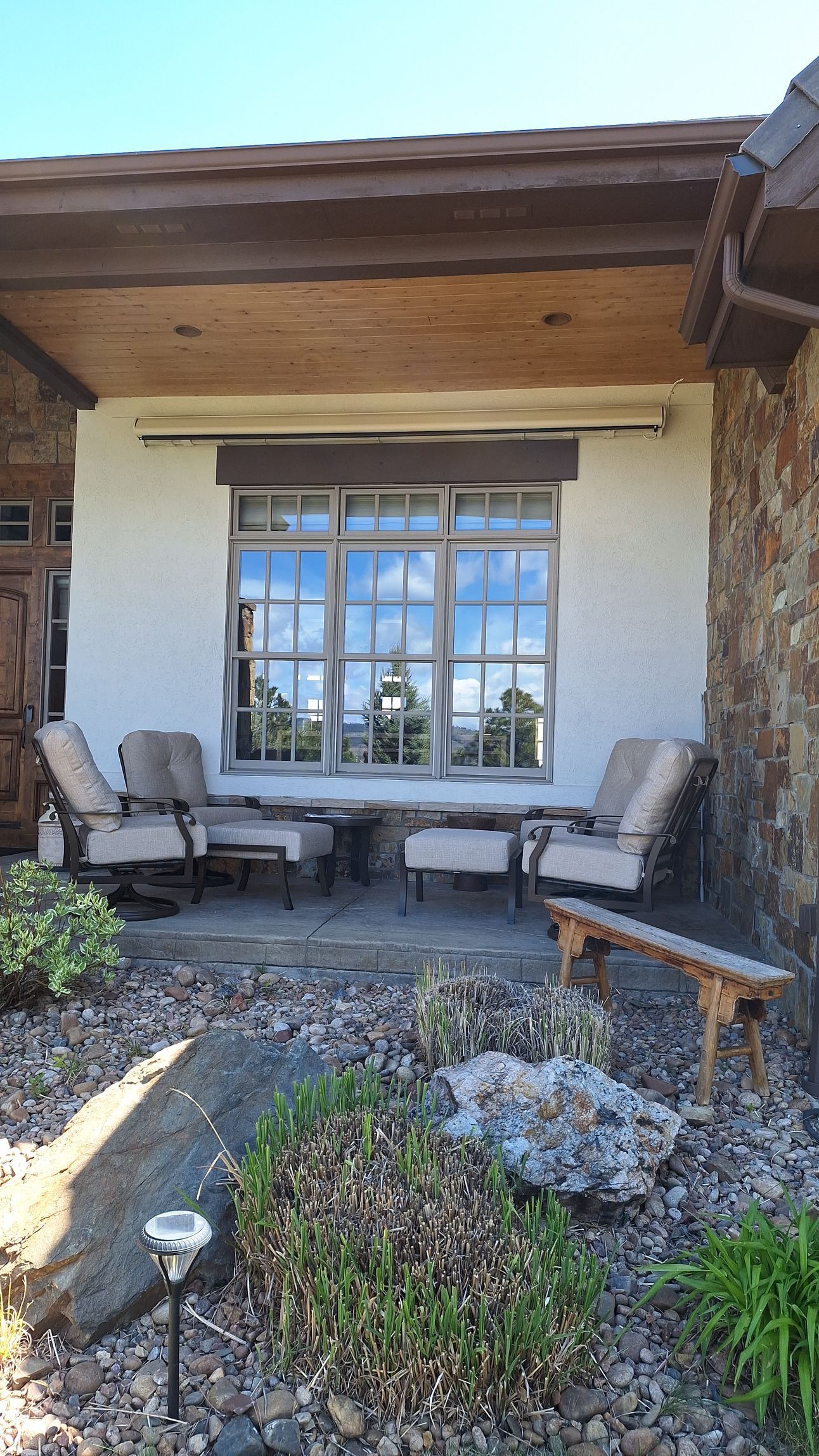 Covered patio with two white chairs and a table beside a large window and stone planter beds.