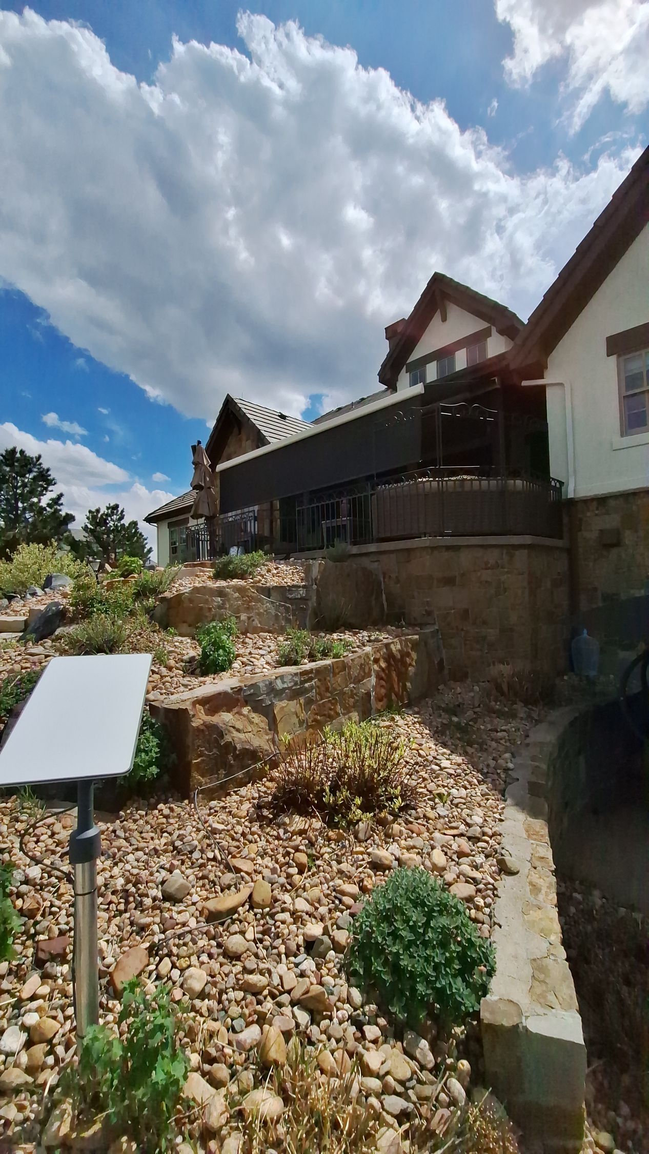 Rocky garden slope beside a house with a dark wooden deck under a partly cloudy sky