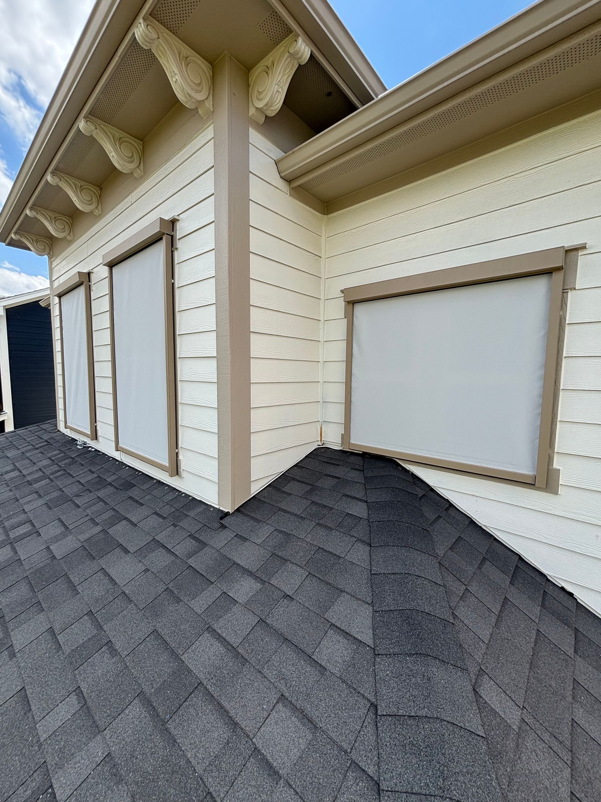 Exterior corner of a beige house with white trim and dark gray shingled roof, showing shuttered windows