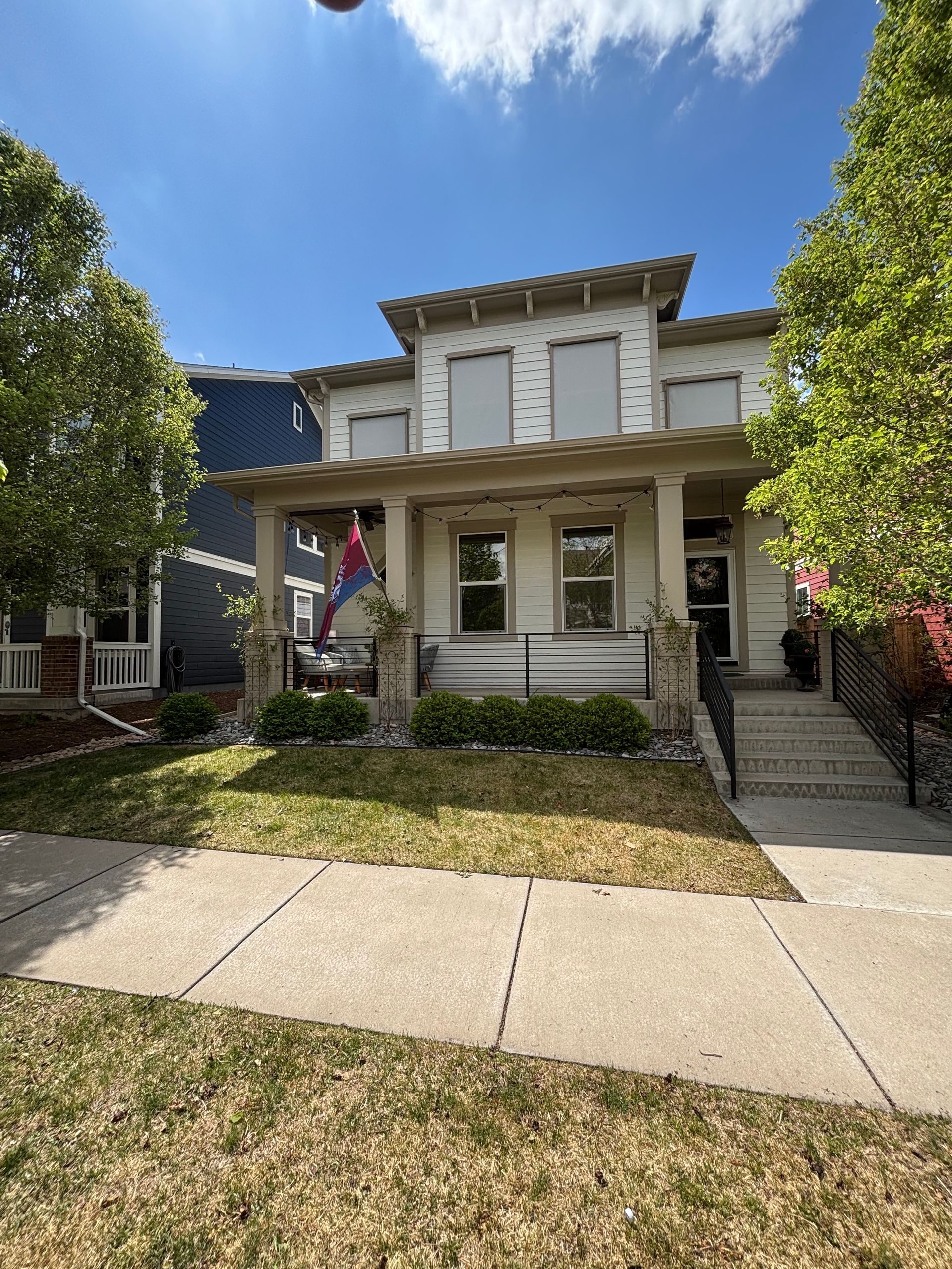 Front view of a gray two-story house with a porch, flag, and tree-lined sidewalk under a blue sky