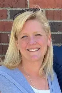 Woman with blonde hair smiles broadly, wearing a light blue sweater and a necklace, in front of a brick wall.
