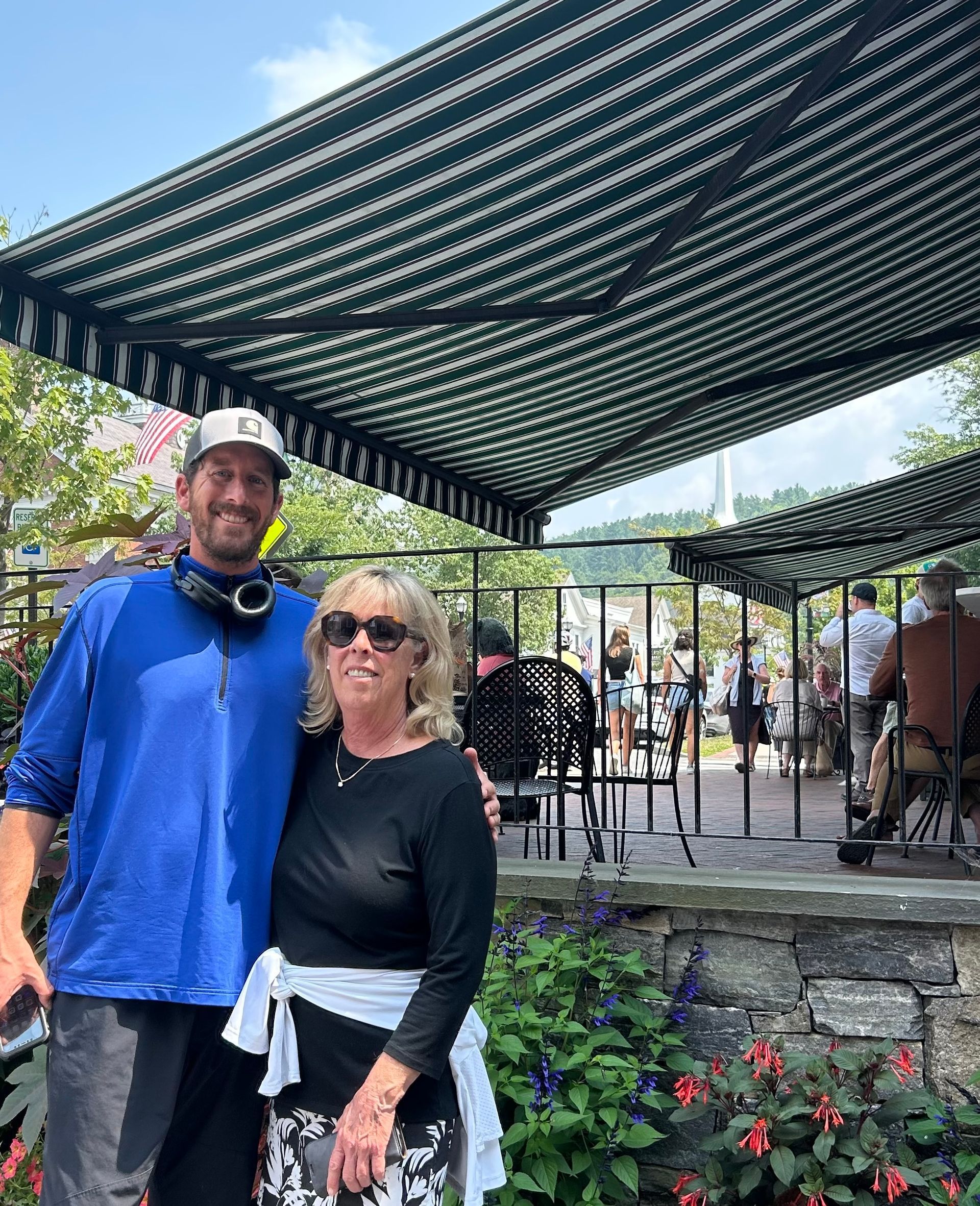 Two people smiling under a shaded pergola in a garden with flowers and stone planters