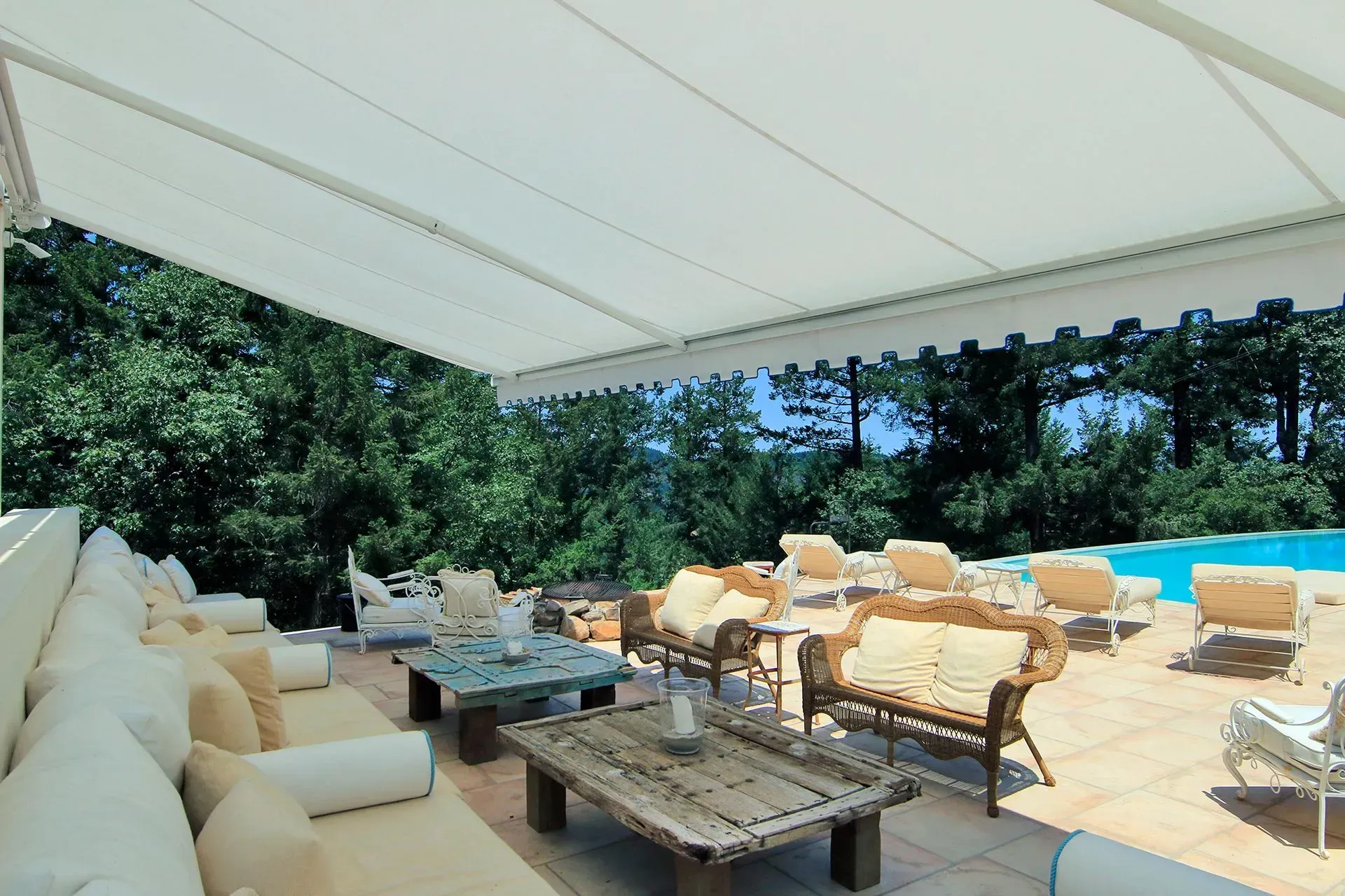 Covered patio with white outdoor seating, stone coffee tables, and a pool beyond trees and blue sky