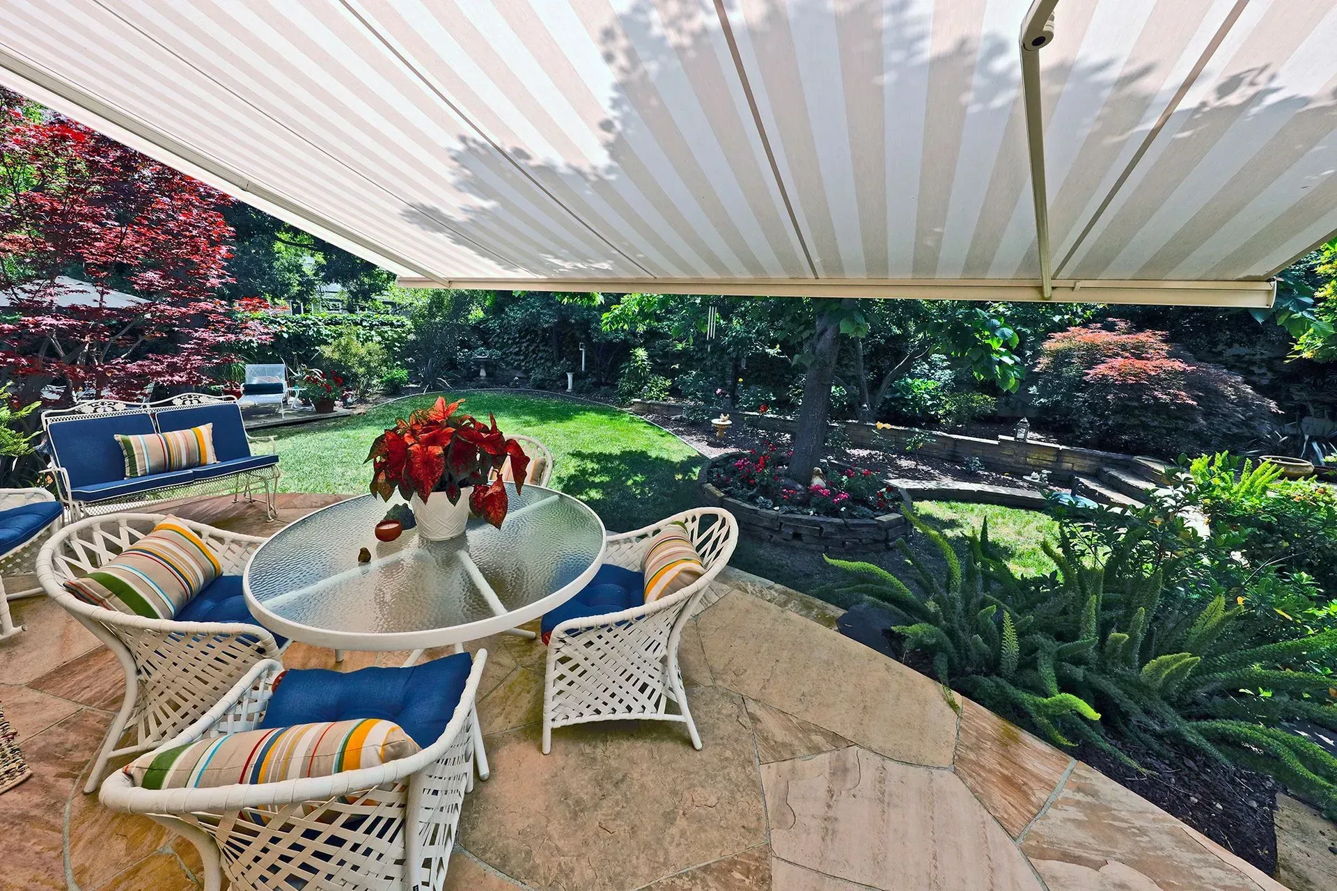 Outdoor patio with white wicker chairs, glass table, and red flowers under a striped awning, overlooking a lush garden