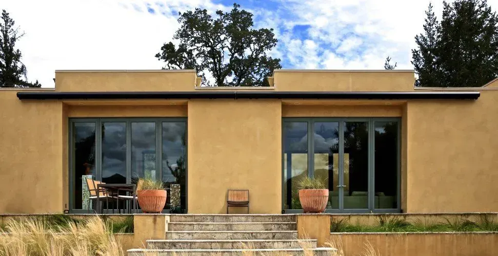 Modern beige house facade with large black-framed windows and a central stone steps entrance, flanked by potted plants