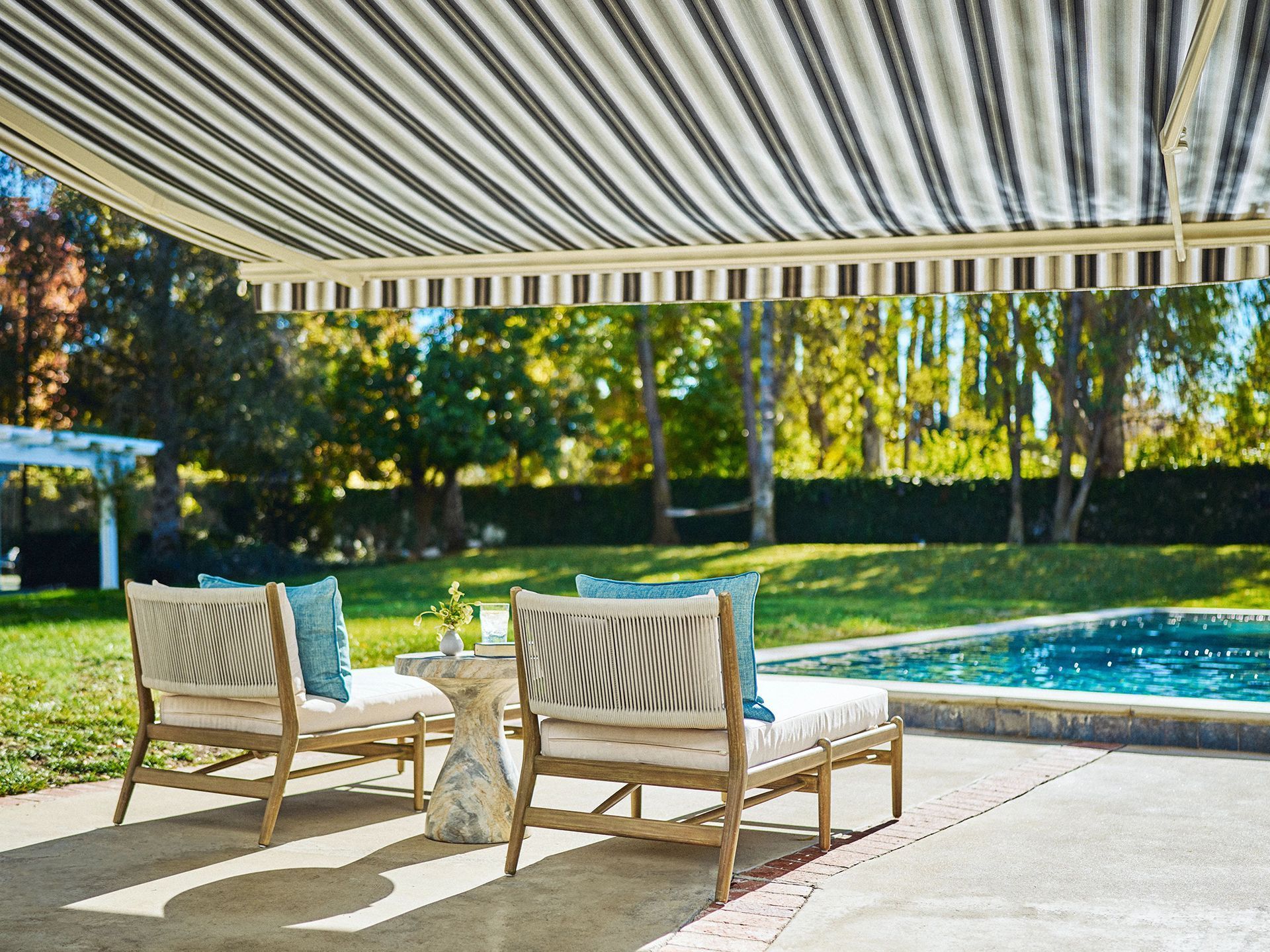 Patio with striped awning, lounge chairs, and pool in a sunny backyard.
