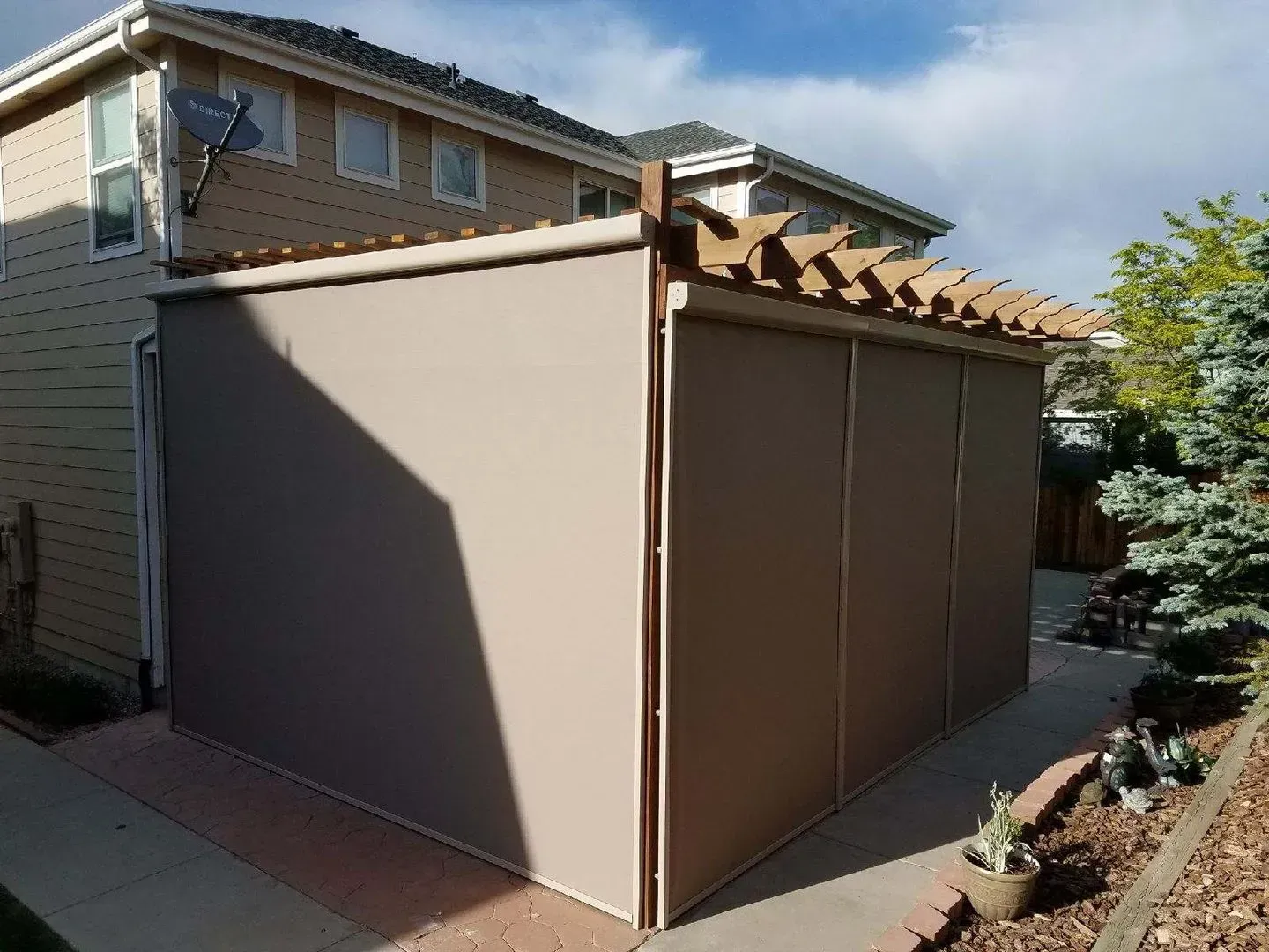 Brown outdoor shed attached to a house, with a slatted roof and plain side panels in a backyard.