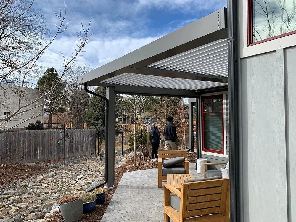 Backyard patio with a slatted pergola, potted plants, and two people walking along a narrow path