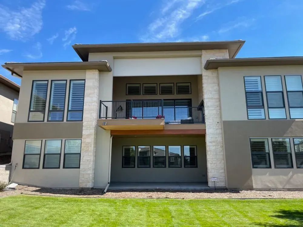 Back of a modern two-story home with a tan stucco exterior, windows, a balcony, and a green lawn under a blue sky.