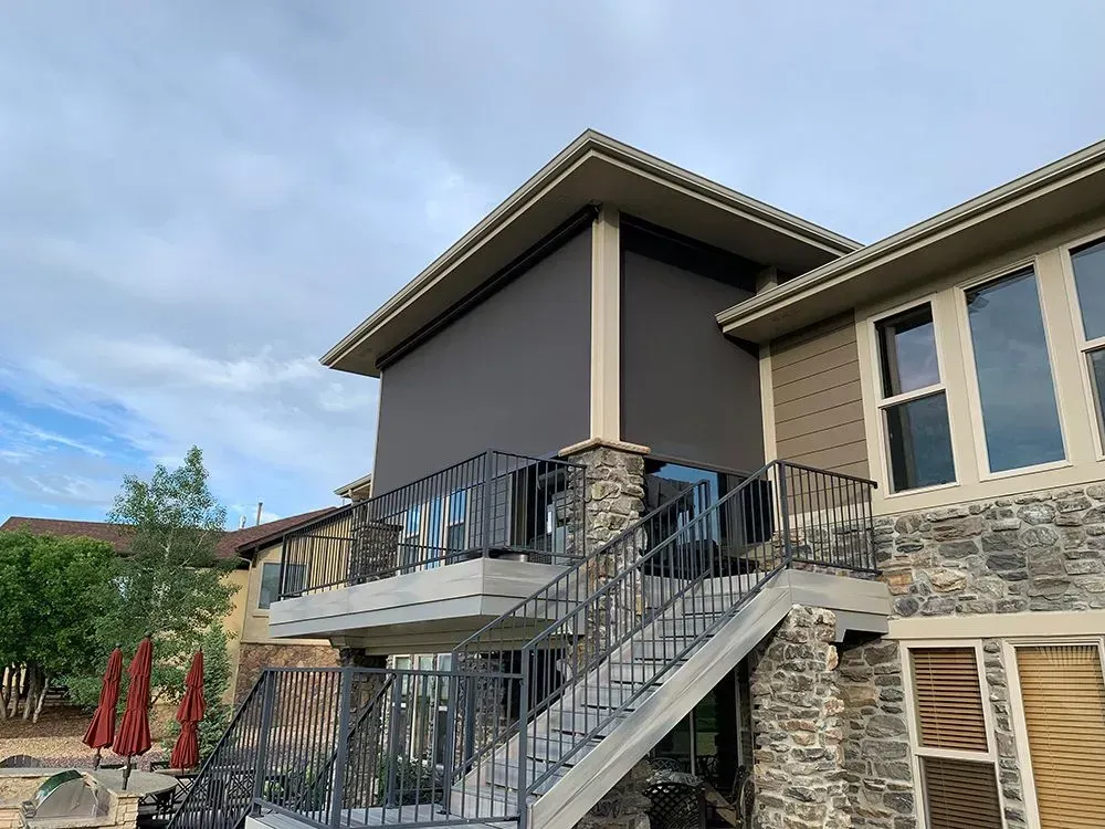 A modern house with stone and tan siding, featuring a covered deck with retractable shade and stairs.