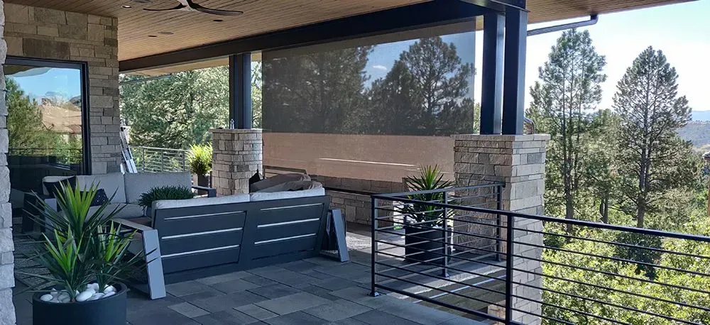 Covered patio with seating, plants, and a view of trees. Black railings and stone columns.