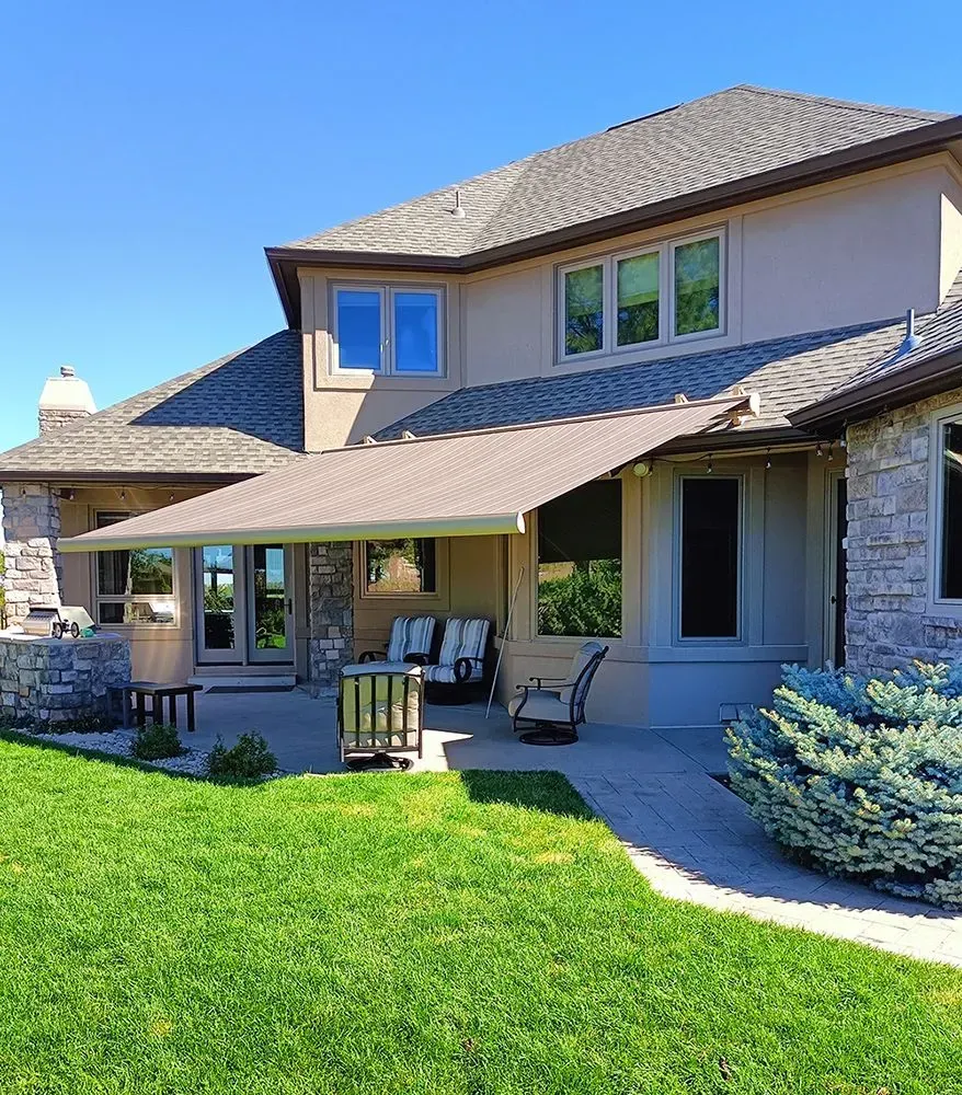 Back patio of a two-story beige house with a tan awning, green lawn, and blue sky.