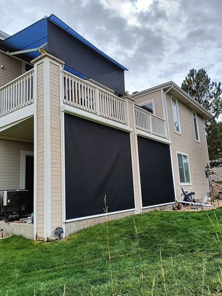Two-story house with black retractable shades on a deck. Green lawn in front, overcast sky.