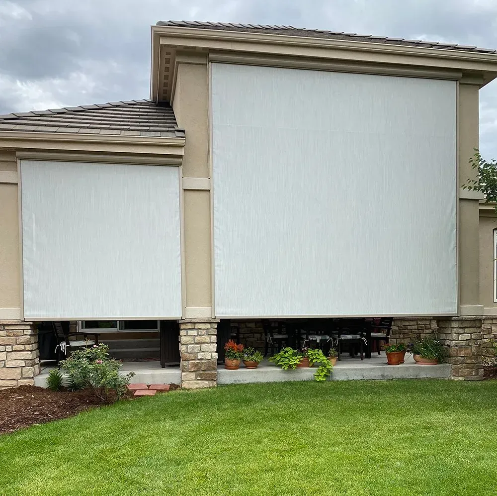 White retractable outdoor sun shades on a house with a stone porch. Green lawn and plants in foreground.