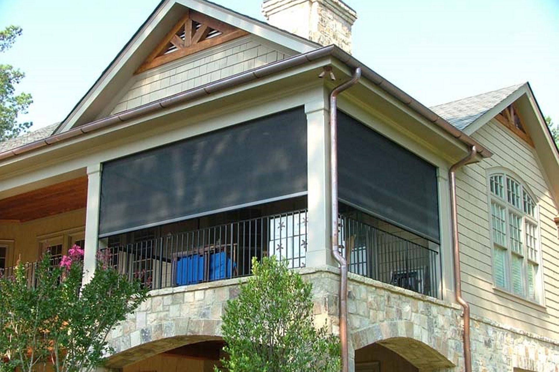 Porch with stone facade, covered by dark retractable shades, attached to a light-colored house.