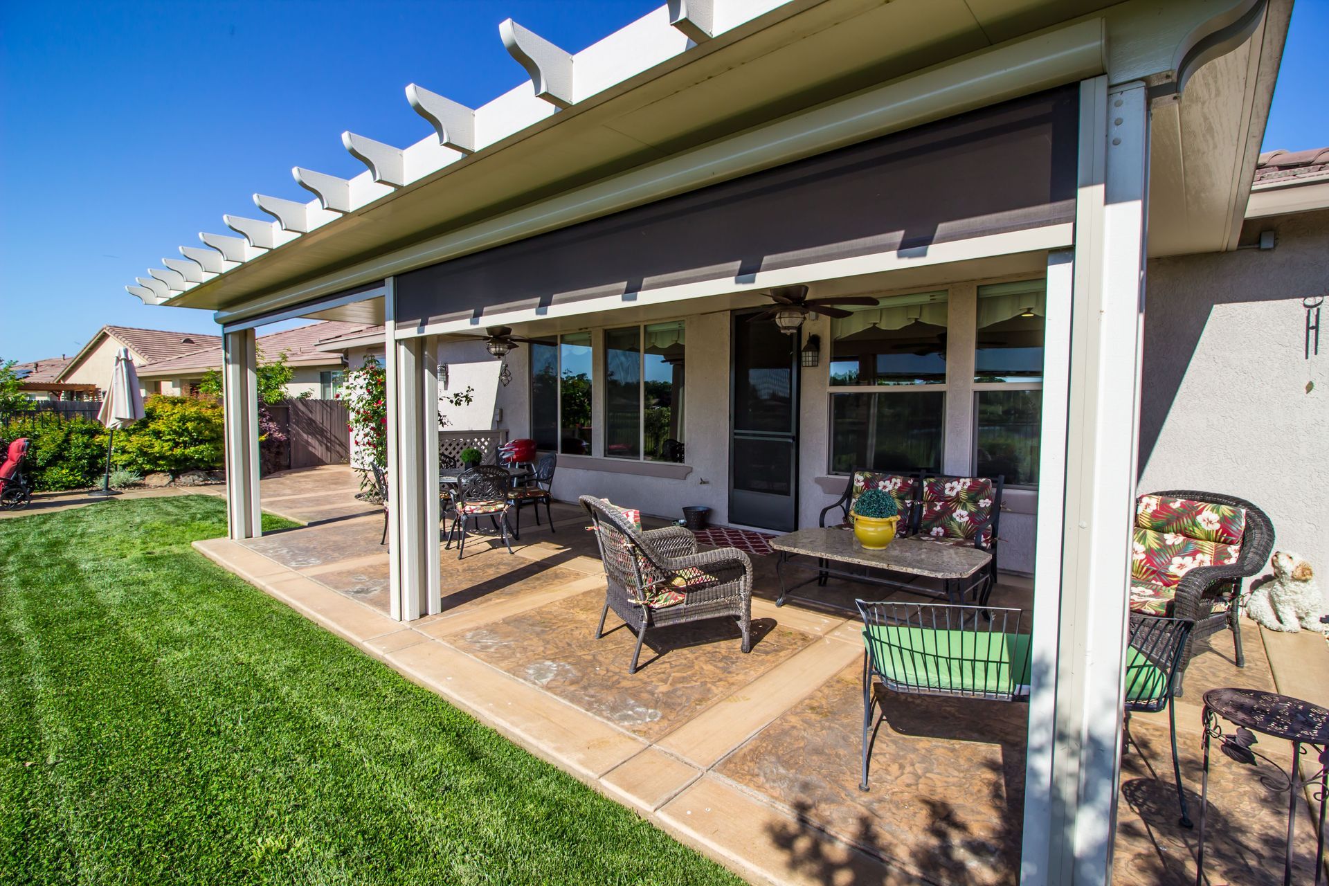 Patio with outdoor chairs and table beside a house, with a green lawn and covered pergola.