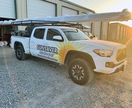 White pickup truck with long roof rack and SUNSAVER logo parked on gravel beside industrial buildings at sunset