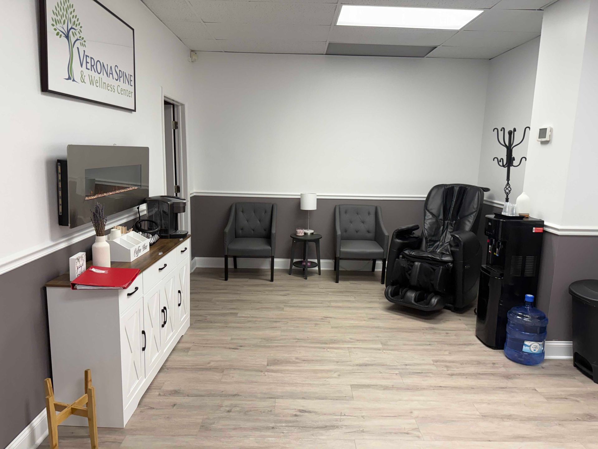 Small waiting room with TV, chairs, black recliner, white cabinet, and water cooler.