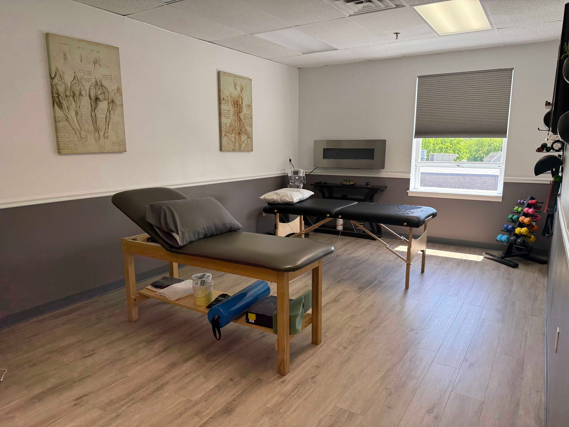 Massage therapy room with table, pillows, wall art, chairs, and a window with blinds