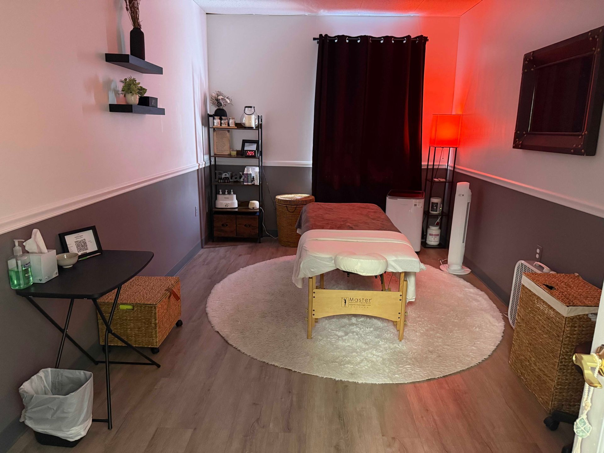 Massage room with treatment table, round rug, dark curtain, shelves, and warm red lighting.