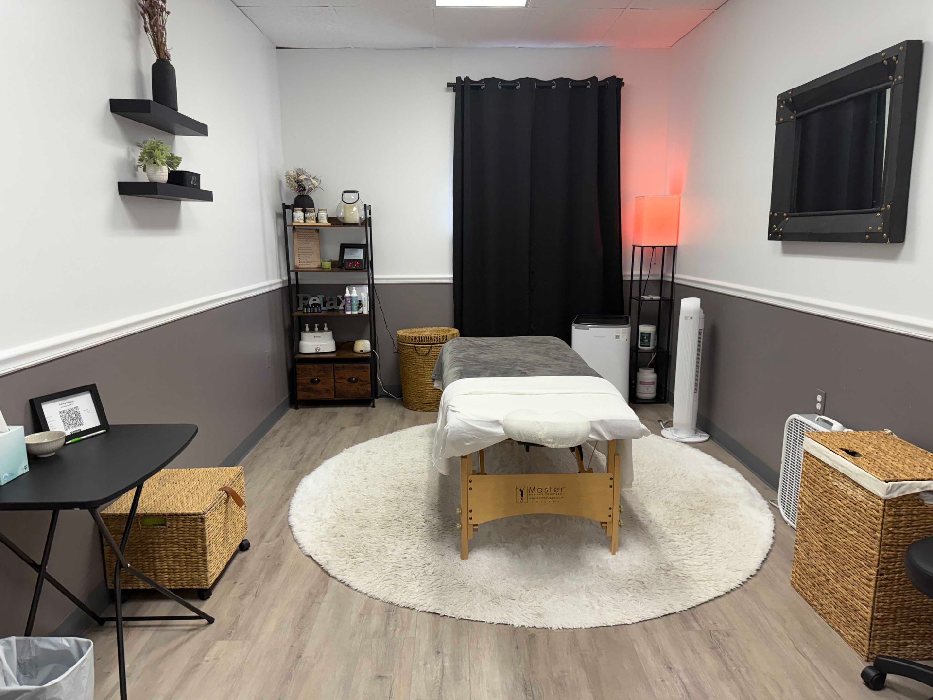 Massage room with table, round rug, black curtains, shelves, and wicker storage baskets.