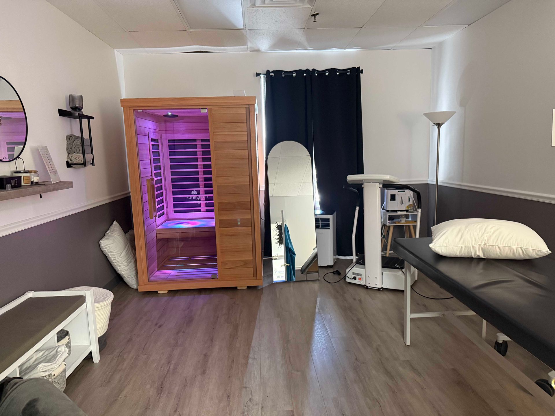 Small therapy room with a purple-lit sauna, treatment bed, white bench, mirror, and black privacy curtain.