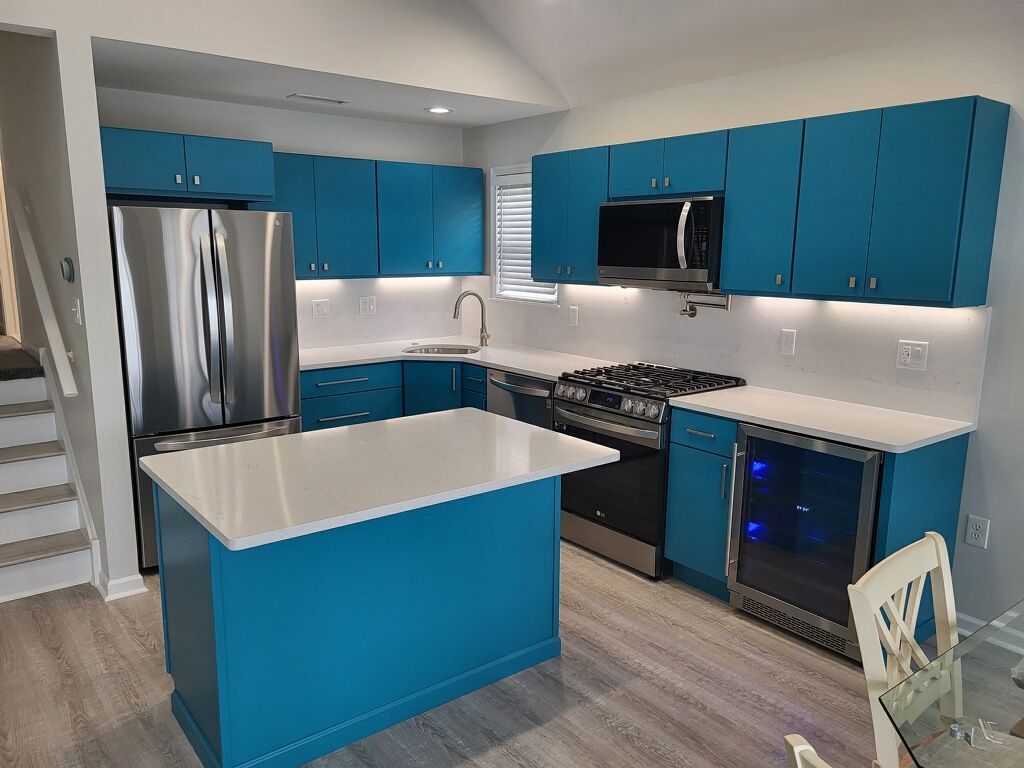 A modern kitchen with turquoise cabinets, white countertops, and stainless steel appliances. An island is in the center, and a wine cooler is visible.