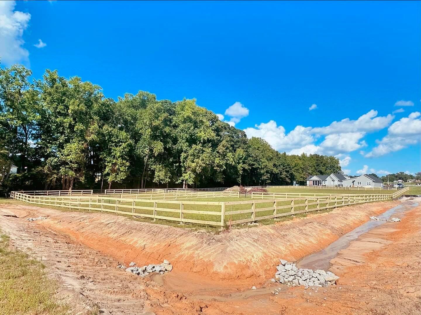 A wooden fence surrounds a dirt field with trees in the background.