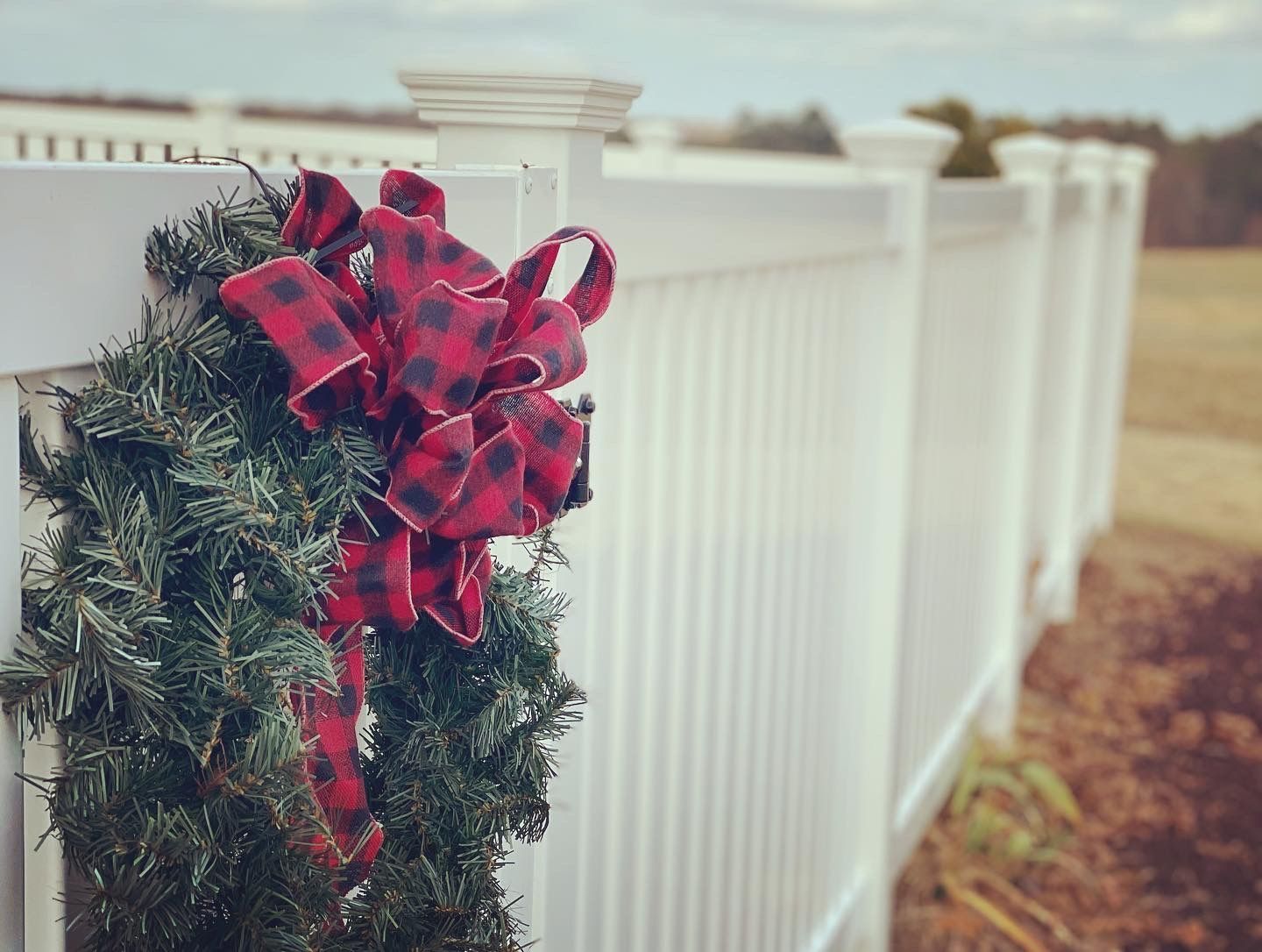 A christmas wreath with a red and black plaid bow is hanging on a white fence.