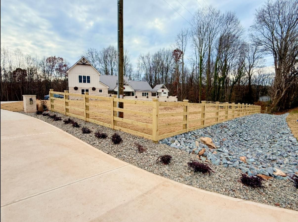 Wooden fence surrounds gravel and bushes near a driveway and house on a cloudy day.