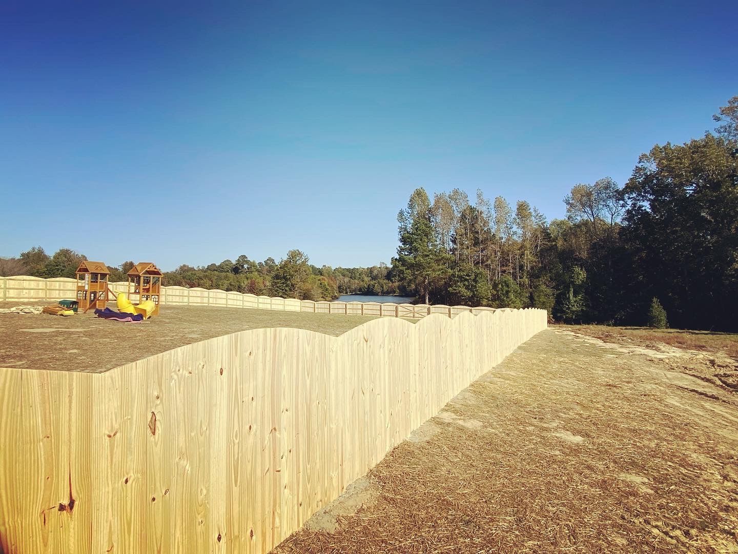 A wooden fence surrounds a playground with a lake in the background.