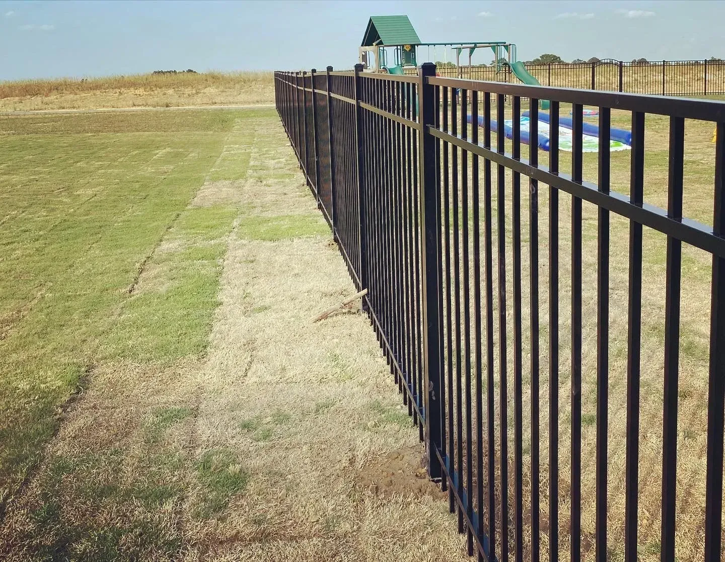 A black fence surrounds a grassy field with a swing set in the background.