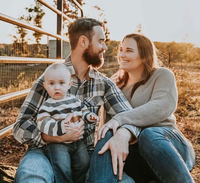Family sitting together outdoors in warm sunset light, smiling beside a fence.