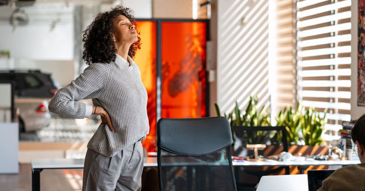 Person stretching with hands on hips in a bright home office kitchen near a sunlit window.