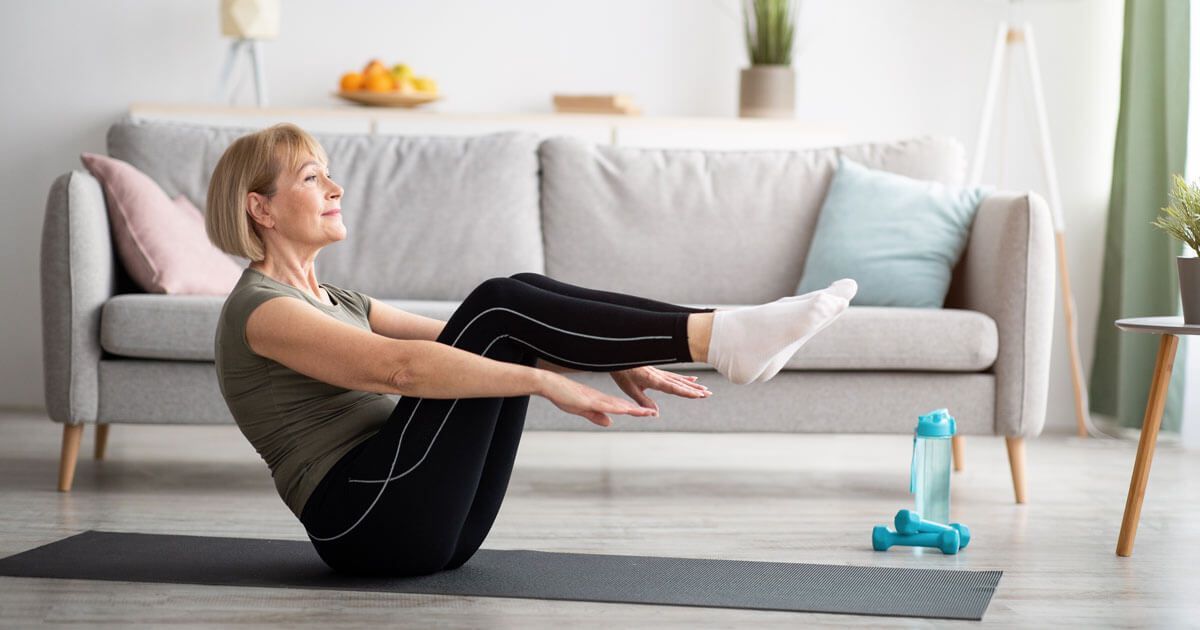 Woman doing seated core exercise on a yoga mat in a bright living room with dumbbells nearby