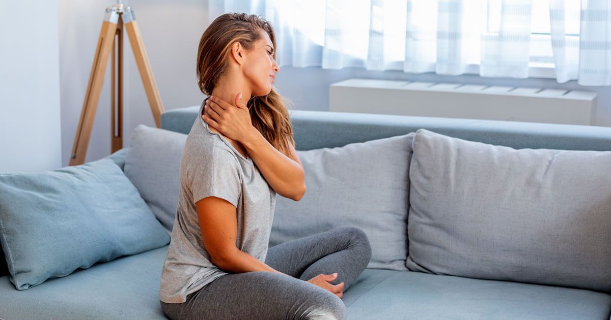 Woman sitting on a couch, holding her neck and looking pained in a bright living room.
