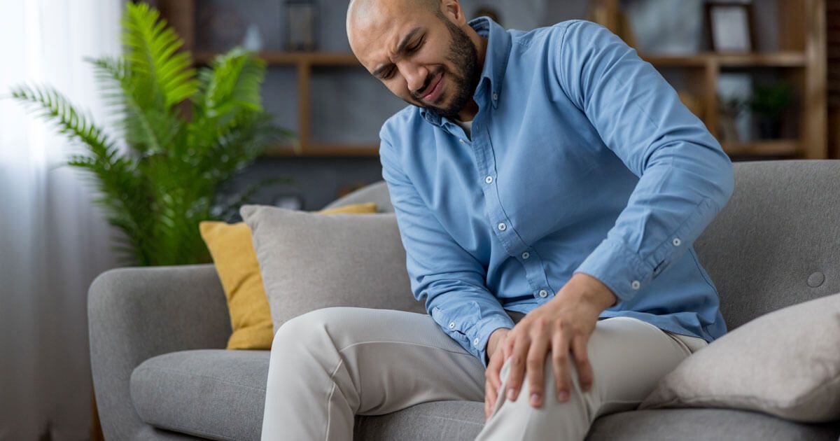 Man sitting on a gray sofa, holding his knee in pain in a living room