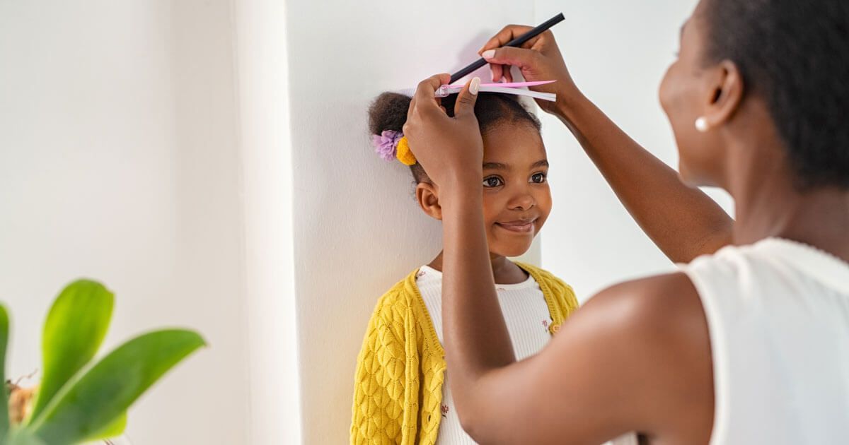Woman combs a child’s hair indoors beside a white wall and a green plant