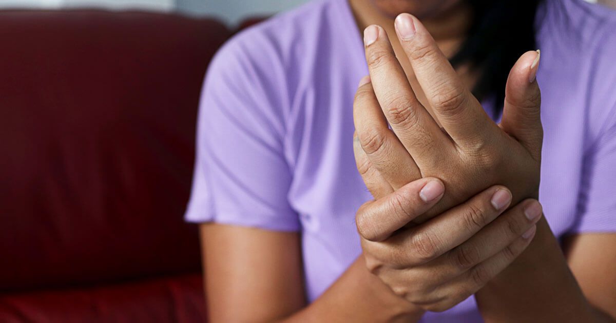 Person holding their wrist in pain while sitting on a red couch, wearing a purple shirt.