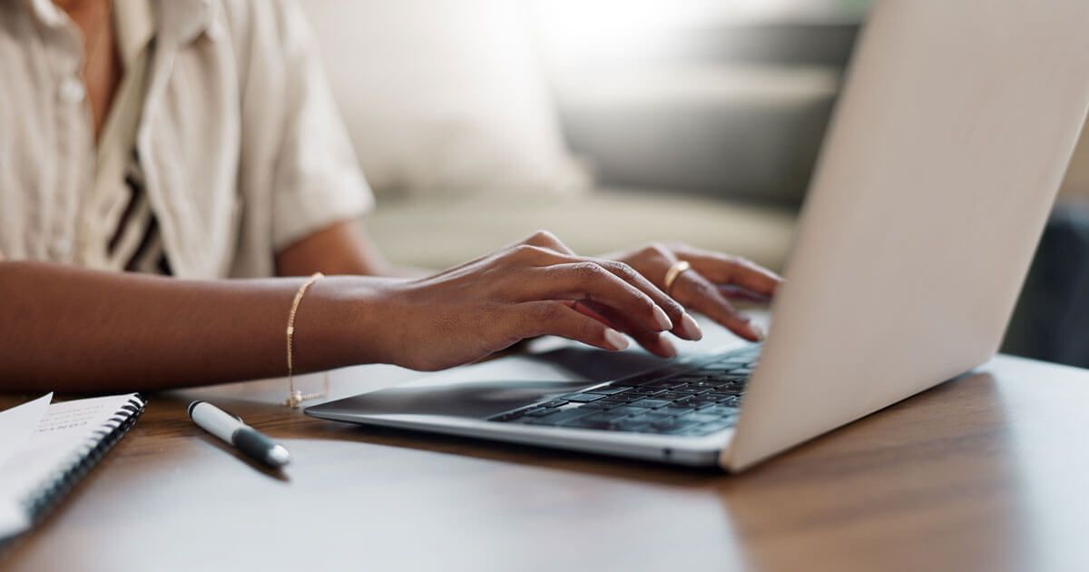 Person typing on a laptop at a desk with a notebook and pen nearby