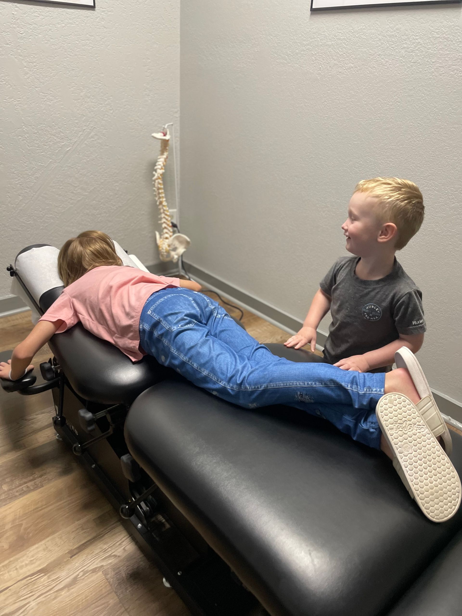 Two children on a padded therapy table, one lying face down and the other sitting nearby and smiling.