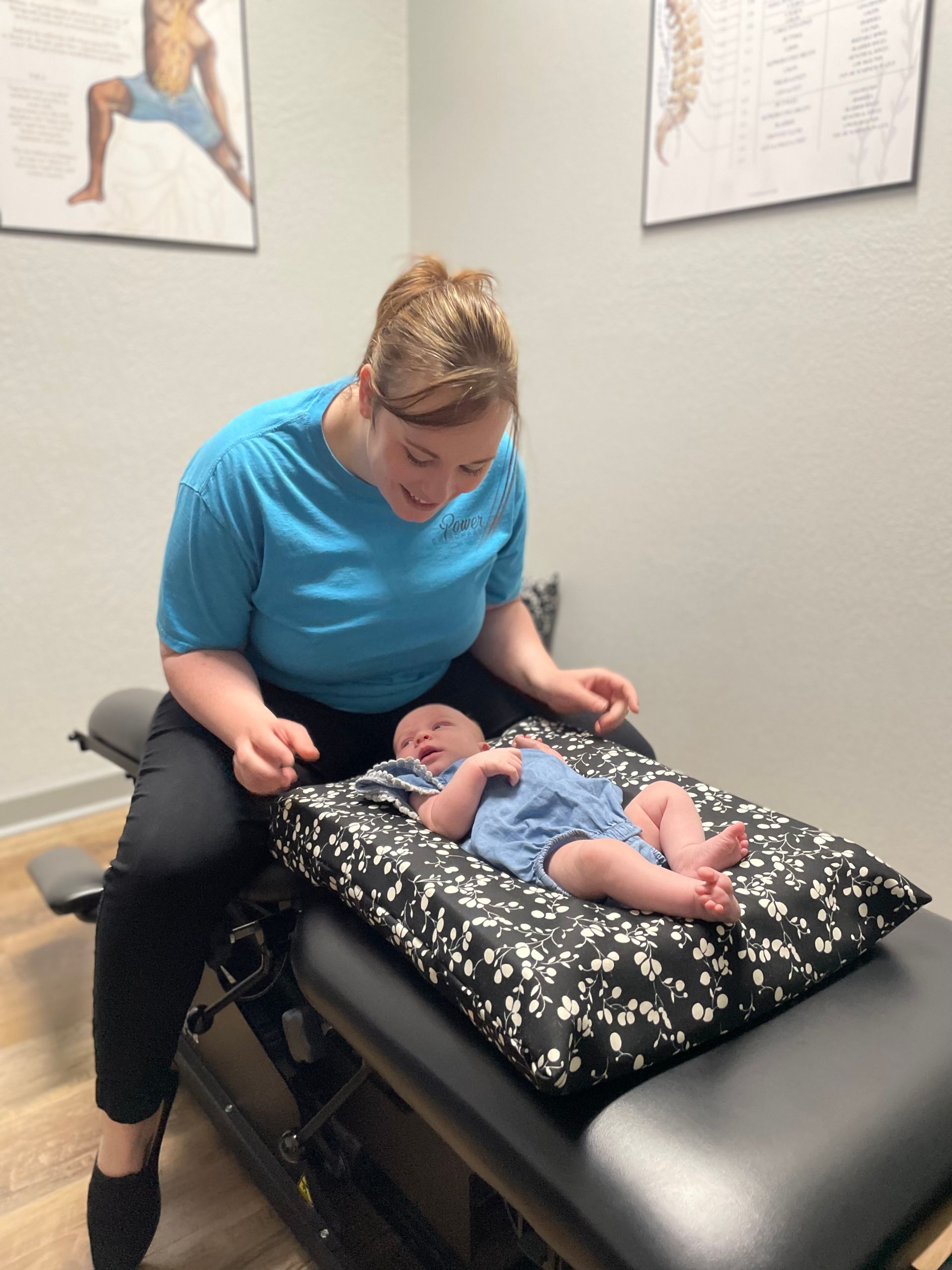 Woman in blue shirt gently examines a baby on a black padded therapy table.